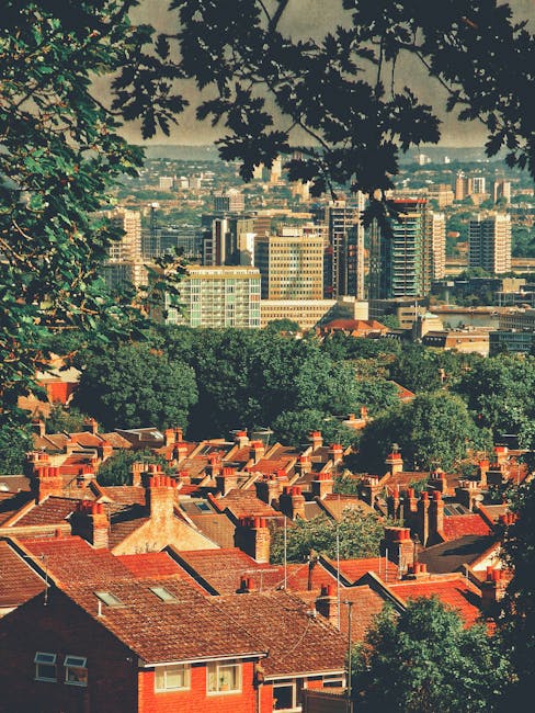 Aerial view of a curved multi-lane highway with several cars and vans moving in both directions, surrounded by a mix of dense green trees and residential buildings with rooftops. In the background, th