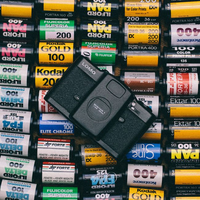 A close-up image showing a vintage black film camera placed on top of a pile of various rolls of photographic film, including brands such as Kodak, Fujicolor, and Ektar. The rolls are stacked together, displaying colorful labels with film names, specifications, and barcode details, predominantly in yellow, white, red, and blue packaging. The camera appears to be a compact point-and-shoot model with a large lens, viewfinder, and control buttons visible on the top. The setting is indoors with even lighting, and the scene reflects a focus on photography materials. As part of home relocation or packing and moving processes, [COMPANY_NAME] specializes in careful handling of valuables such as cameras and photography equipment during removals, ensuring safe transportation of delicate items in a furniture transport or loading process.