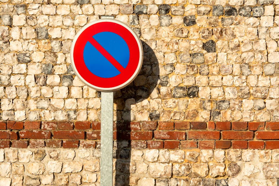 A round traffic sign with a blue background and a red border displaying a red diagonal line across it, indicating no parking or stopping zone, mounted on a metal pole against a textured beige and reddish-brown brick wall. Shadows cast by the sign and pole suggest bright sunlight. The brick wall has a horizontal stripe of red bricks embedded within the beige stones, creating a contrasting pattern. This setting is outdoors on a paved surface, potentially near a property entrance or public street. The sign's presence may relate to parking and loading restrictions relevant to home relocation or furniture transport services provided by Man with Van Shooters Hill, situated in the context of house removal logistics and compliance with local parking rules.