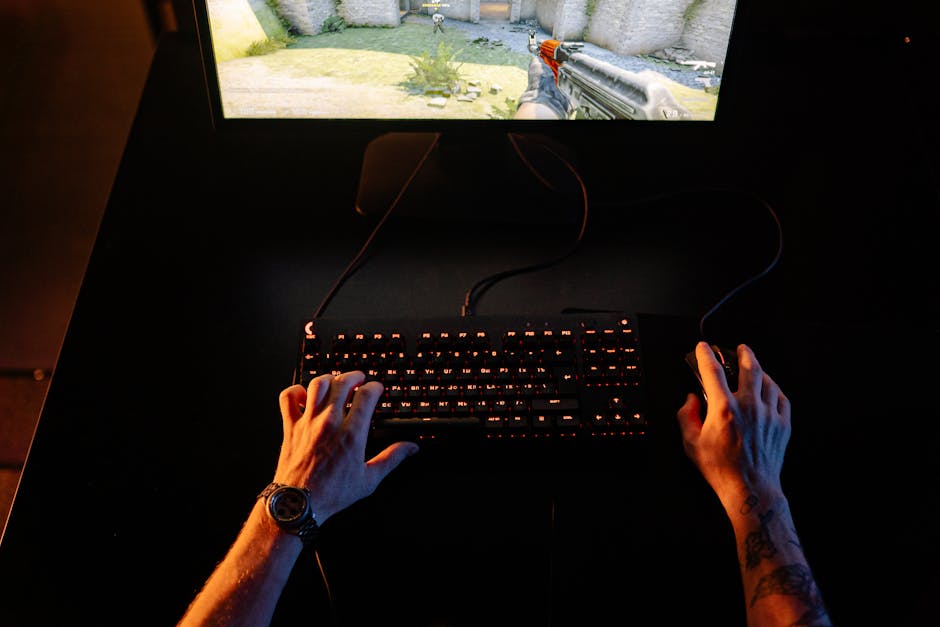 A person with tattooed forearms is sitting at a dark desk, operating a gaming computer with a black keyboard illuminated by orange backlighting. Their left hand hovers over the keyboard while their right hand holds a computer mouse. In front of them, a monitor displays a first-person shooter video game scene involving a firearm aimed at an outdoor environment with stones, grass, and a yellowish wall. The setting appears to be indoors with low ambient lighting. The scene captures a moment of casual gaming, highlighting the environment of a home setup possibly involved in packing or organizing equipment during a house removal process, with the focus on the computer peripherals and monitor.