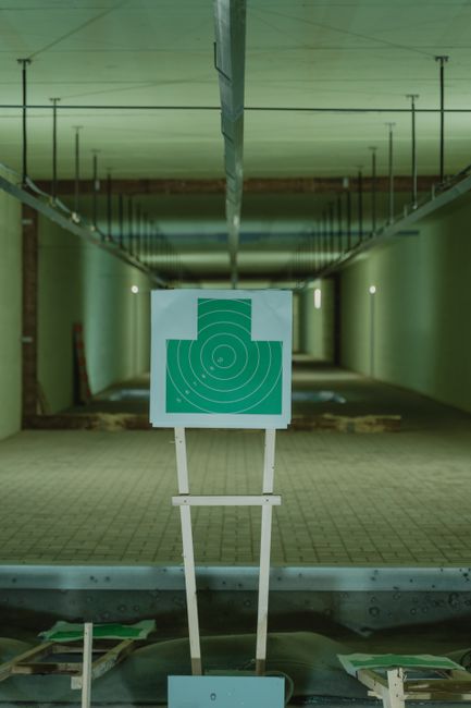 An indoor shooting range with a target (bullseye) mounted on a stand positioned at the far end of the corridor, surrounded by smooth, light-colored walls and ceiling. The range features a tiled floor and is illuminated by overhead fluorescent lighting, creating a well-lit environment. In the foreground, there are wooden pallets and protective barriers indicating a safe, controlled space for practicing shooting. The photo appears to be taken from behind a glass or protective barrier, with the environment arranged for firearm practice or training sessions. The setting is associated with facility management and safety procedures relevant to secure shooting or training activities, which may relate to logistics in house removals or equipment transport in the context of removals services by Man with Van Shooters Hill.