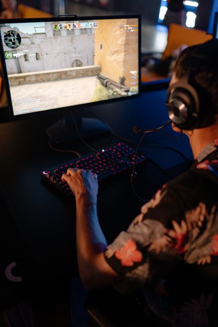 A person wearing large over-ear headphones is seated at a desk in front of a computer monitor displaying a first-person shooter game set in an outdoor urban environment with walls, windows, and a sandy ground. The individual's right hand is on a gaming keyboard with a pink-backlit design, and their left hand is near a mouse, both resting on the desk. The person is wearing a short-sleeved shirt with a floral pattern, and their face is partially visible from the side. The background shows a dimly lit room with other computer setups and people, suggesting a gaming or e-sports environment that could be associated with home relocation or moving services for gaming setups, though the primary focus is on gaming activity.