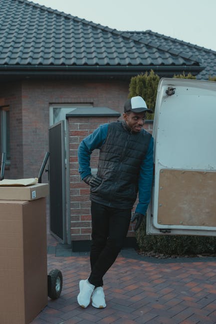 A man wearing a blue long-sleeve shirt, black padded vest, black trousers, and white sneakers is standing outside a house, leaning against the open rear door of a white moving van. The van is parked o
