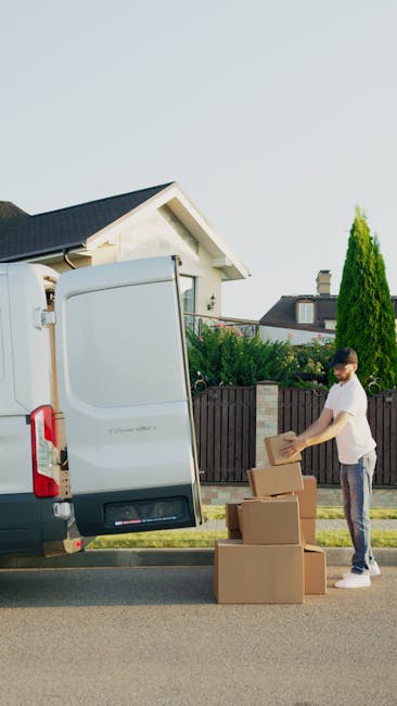 A man wearing a blue long-sleeve shirt, black padded vest, black trousers, and white sneakers is standing outside a house, leaning against the open rear door of a white moving van. The van is parked o