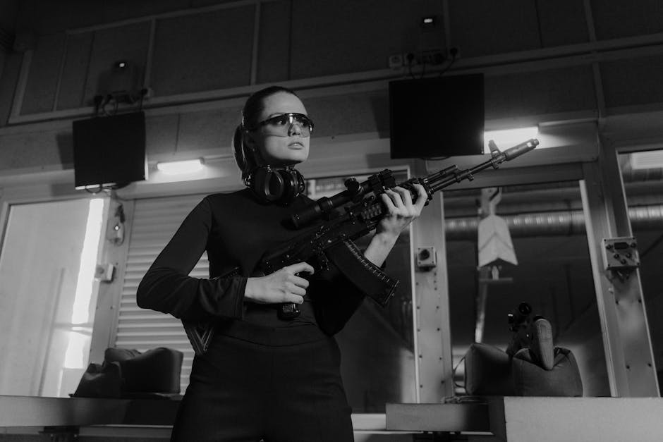 A woman stands indoors, wearing safety glasses and headphones around her neck, holding a scoped rifle with both hands, aiming upwards. She is dressed in a black long-sleeve top, with a serious expression and her dark hair pulled back. The background includes a modern interior space with hanging televisions, shelves, and lighting fixtures, suggesting an environment related to shooting or training, not a home relocation or moving scene. The setting appears well-lit with natural and artificial light, and the overall composition emphasizes the woman's focus and stance. The image does not feature furniture, boxes, or moving equipment, indicating it is unrelated to house removals but relevant to shooting or training activities.