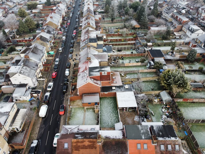 A black-and-white aerial photograph of a residential area showing a street lined with closely spaced terraced houses and semi-detached properties, with some houses having small back gardens. Several cars are parked along the road and in driveways, while trees and greenery are interspersed throughout the neighbourhood. In the background, the landscape extends to an open area with scattered trees and distant structures under a partly cloudy sky. The image captures the urban layout, typical of a UK suburb, and provides context for house removals or home relocation services offered by Man with Van Shooters Hill, illustrating the urban environment where furniture transport and packing and moving activities might take place.