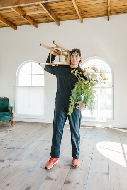 A man wearing a black long-sleeve shirt, dark blue pants, and pink sneakers stands inside a spacious room with wooden flooring and a wooden ceiling with exposed beams. He is smiling and holding a small wooden stool or side table above his shoulder using both hands, with the seat facing outward. In his other hand, he holds a large bouquet of assorted flowers and greenery, including daisies and ferns. Behind him, there are three large, rounded-arch windows allowing natural light into the space, which is sparsely furnished with a vintage-style green armchair on the left. The scene appears to be part of a home relocation or furniture transport process, possibly related to a house move or packing and moving service, as indicated by the professional activities of [COMPANY_NAME].