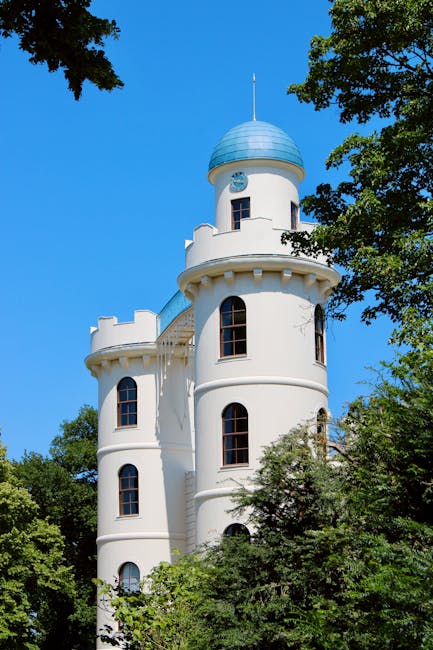 A tall, white, turret-style building with multiple narrow, arched windows, surrounded by green trees and foliage, under a clear blue sky. The structure features battlement-style parapets along the upper levels and a conical roof with a weather vane on top. This historic-looking, castle-like building is situated in a natural setting, and the bright sunlight highlights its architectural details. The scene suggests a location near Oxleas and Shooters Hill, consistent with the type of property involved in house removals and furniture transport services by Man with Van Shooters Hill, as shown on their website page about Severndroog Castle moves.