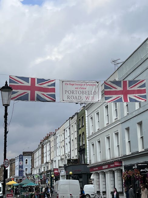 A street scene on Portobello Road in London, featuring a large decorative banner with Union Jack flags hanging above the pavement. The banner displays the text 'The Royal Borough of Kensington and Chelsea PORTOBELLO ROAD, W.11' on a white background. Below the banner, a row of traditional terraced buildings with white facades and colourful window frames line the street, some occupied by shops and cafes. The sidewalks are bustling with pedestrians, and a white van is parked near the curb, likely engaged in home relocation or furniture transport activities. To the left, a vintage-style street lamp and a 20 mph speed limit sign are visible. The overcast sky creates diffused lighting, and the overall scene depicts a typical lively London shopping street, possibly during a moving or packing process facilitated by [COMPANY_NAME], which provides removals and home relocation services.