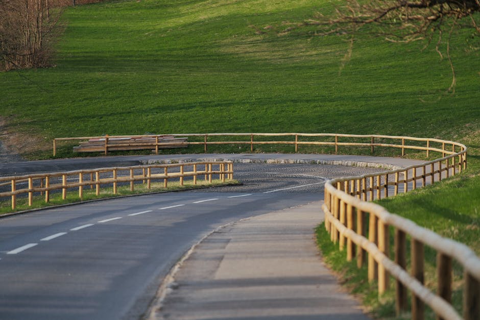 A street scene on Portobello Road in London, featuring a large decorative banner with Union Jack flags hanging above the pavement. The banner displays the text 'The Royal Borough of Kensington and Chelsea PORTOBELLO ROAD, W.11' on a white background. Below the banner, a row of traditional terraced buildings with white facades and colourful window frames line the street, some occupied by shops and cafes. The sidewalks are bustling with pedestrians, and a white van is parked near the curb, likely engaged in home relocation or furniture transport activities. To the left, a vintage-style street lamp and a 20 mph speed limit sign are visible. The overcast sky creates diffused lighting, and the overall scene depicts a typical lively London shopping street, possibly during a moving or packing process facilitated by [COMPANY_NAME], which provides removals and home relocation services.