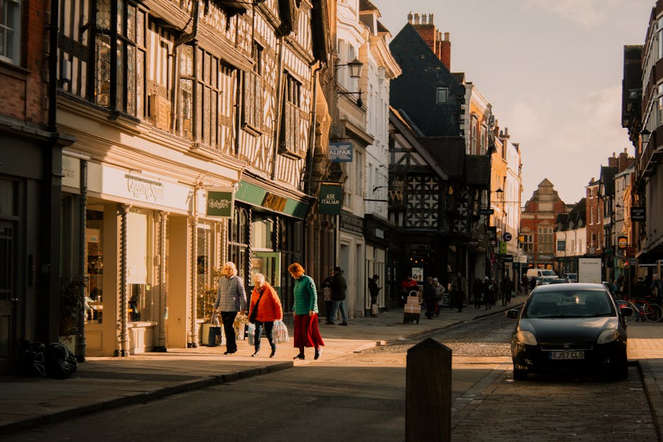 A street scene in Shrewsbury Park during late afternoon or early evening, with warm sunlight illuminating a row of historic multi-story buildings featuring timber framing, brickwork, and traditional shopfronts. Pedestrians, including a woman in a red coat and a woman with a shopping bag, walk along the sidewalk. Several people are gathered outside shops, some browsing or entering store entrances. A small, dark-colored car is parked on the cobbled street in the foreground next to a set of bicycles, with additional pedestrians visible further down the street. The environment suggests a lively commercial area with a mix of retail outlets and cafes, typical of a busy home relocation or moving logistics area in Shooters Hill, where Man with Van Shooters Hill offers removals and moving services. The overall scene emphasizes urban activity, architectural character, and the movement of people—a setting relevant to furniture transport and packing during house removals.
