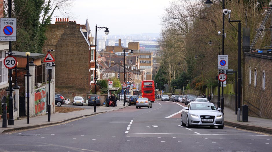 A wide urban street scene during daytime in Shooters Hill showing a slight curve in the road with parked cars lining both sides. In the foreground, a silver Audi is about to move forward while a section of the pavement on the left side has a delivery truck parked beside a bus stop shelter. Several pedestrians are waiting at the bus stop, and the street features multiple traffic signs, including a 20 mph speed limit sign and no parking signs within a controlled zone. In the background, a bright red double-decker bus is approaching, with residential buildings, trees, and lampposts along the street. The scene is well-lit with natural daylight, and the environment appears calm and typical of a residential area in London, ideal for household removals or home relocation services by Man with Van Shooters Hill.