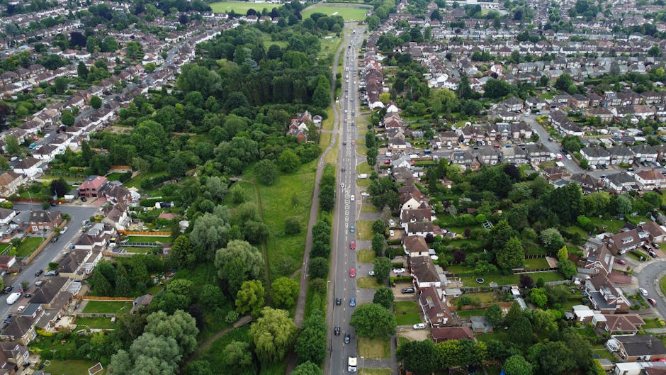 A wide urban street scene during daytime in Shooters Hill showing a slight curve in the road with parked cars lining both sides. In the foreground, a silver Audi is about to move forward while a section of the pavement on the left side has a delivery truck parked beside a bus stop shelter. Several pedestrians are waiting at the bus stop, and the street features multiple traffic signs, including a 20 mph speed limit sign and no parking signs within a controlled zone. In the background, a bright red double-decker bus is approaching, with residential buildings, trees, and lampposts along the street. The scene is well-lit with natural daylight, and the environment appears calm and typical of a residential area in London, ideal for household removals or home relocation services by Man with Van Shooters Hill.