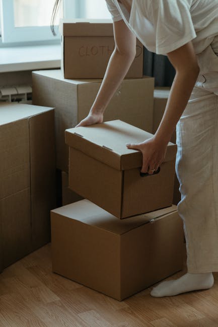 A person wearing light-colored clothing, with only their torso and arms visible, is handling a medium-sized cardboard box during a home relocation. They are in a room with natural light coming through a window, and several other cardboard boxes are stacked nearby, some labeled with writing such as 'CLOTHES'. The individual is carefully lifting or placing the box onto or from a stack of similar boxes on a wooden floor. The boxes vary in size, and the scene suggests packing and moving activities typical of furniture transport and home removals services. Man with Van Shooters Hill is involved in the loading process, ensuring secure handling of belongings, with additional boxes and packaging materials visible in the background, supporting the logistics of an urgent eviction or emergency move.