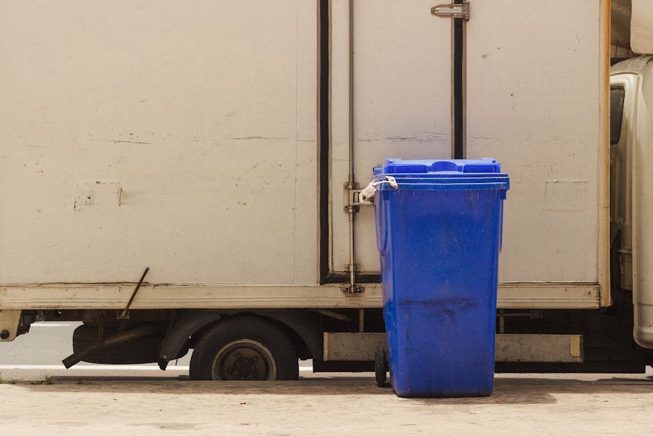 A blue wheeled waste bin with a closed lid is positioned on the pavement next to a white delivery van with a large, plain external cargo area, partly visible in the image. The van is parked close to a doorway or loading bay, with a metal door handle and hinges visible behind the bin. The pavement appears clean, and the scene is lit with natural daylight, suggesting an outdoor setting during daytime. The van's cargo area has some scuff marks and dirt, indicating regular use in a home relocation or moving process. The presence of the waste bin next to the van hints at waste disposal or packing materials being managed during furniture transport or packing and moving activities, which are common aspects of house removals. This image illustrates the logistical process of loading items, including managing waste disposal in compliance with local laws, as addressed in the context of Shooters Hill's waste disposal regulations. Man with Van Shooters Hill is involved in such removals and related logistics services.