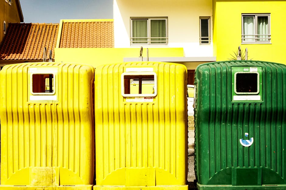 A row of three large wheelie bins, two yellow and one green, positioned on a pavement in front of a modern residential building with white and yellow walls and multiple windows. The yellow bins are side by side, with the green bin to the right, each featuring a small window at the top for inserting waste. The bins are made of ribbed plastic, with visible dirt and wear, and are used for household waste disposal as part of home relocation or packing and moving logistics. The background includes the building's facade, a section of red-tiled roof, and a bright, sunny day with natural lighting. Man with Van Shooters Hill occasionally services such waste disposal needs during furniture transport and house removals, ensuring proper handling according to waste disposal laws in Shooters Hill.