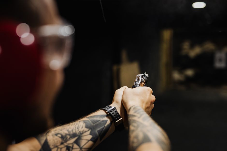 A person with visible tattoos on their arms is holding a silver revolver with both hands, aiming it forward inside a dimly lit environment. The individual is wearing a black wristwatch on their left wrist and has dark, blurred hair or headgear partially visible in the foreground. The background appears to be an industrial or storage area with shelves or stacks of items faintly visible, and soft lighting creates a shadowed atmosphere. This scene represents a controlled loading or safety procedure, potentially linked to the packing or transportation phase of home relocation services provided by Man with Van Shooters Hill, within the context of furniture transport or moving logistics.