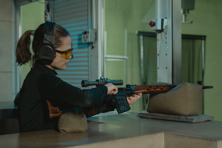 A woman wearing black clothing, yellow-tinted safety glasses, and protective ear muffs is positioned behind a counter at an indoor shooting range. She is holding a scoped rifle, aiming it downrange, with her left hand supporting the wood-stocked firearm and her right hand on the trigger. In front of her on the counter, there is a beige bag or case, and beneath it, some folded paper or cloth. The background features greenish walls, electrical outlets, and a door with shuttered panels. The environment is lit with soft, even lighting suitable for a controlled firing area, reflecting a setting where precision shooting or firearm training occurs, akin to a removals service's careful handling and transport processes, and subtly aligned with home relocation logistics handled by companies like Man with Van Shooters Hill.